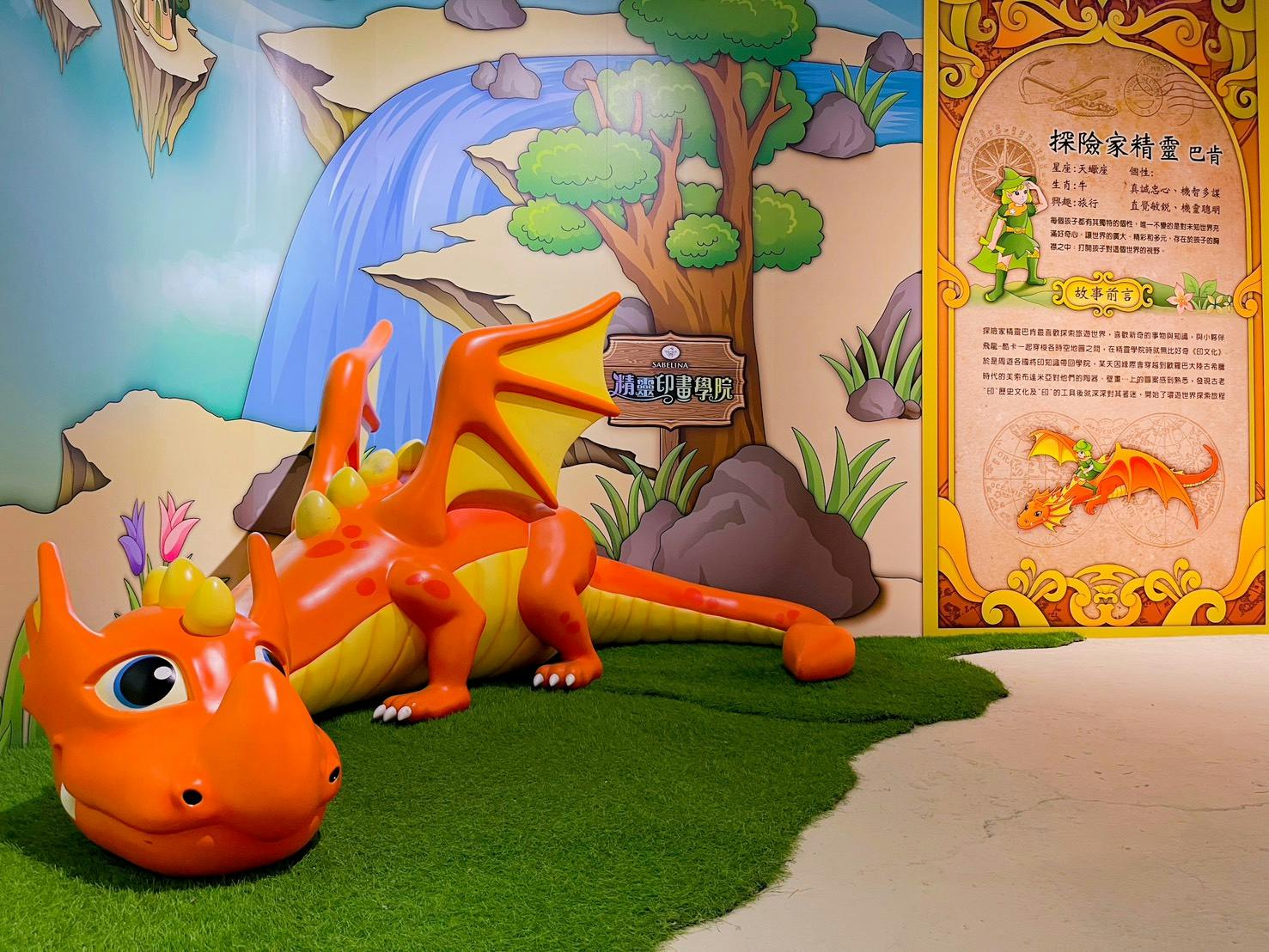 A whimsical orange dragon sculpture on green turf in front of a colorful mural with a waterfall and trees.