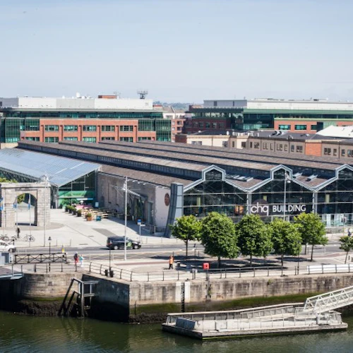 En urban scen vid floden med moderna och historiska byggnader, träd och en dockad brygga, under en klar himmel.