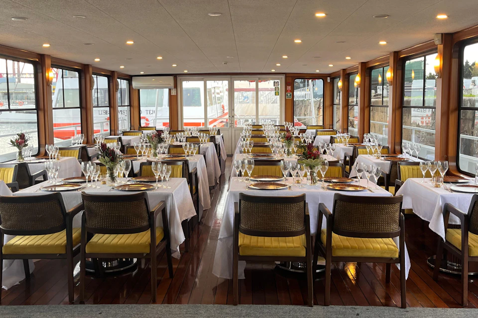 A neatly set dining area with white tablecloths, yellow chairs, and flower centerpieces, located in a boat with large windows.