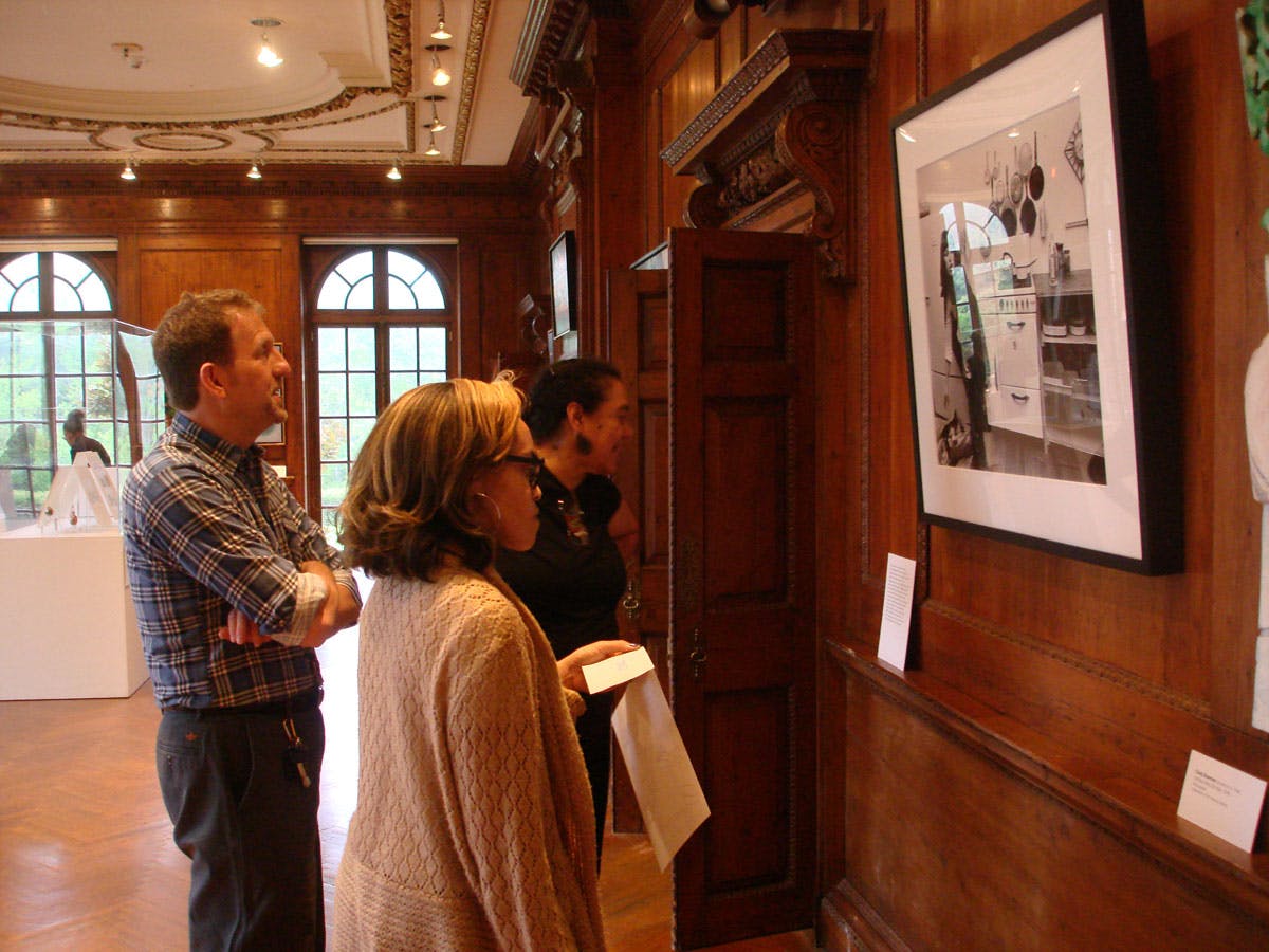 Tre personer observerer indrammet kunst i et galleri med træpaneler med buede vinduer i baggrunden.