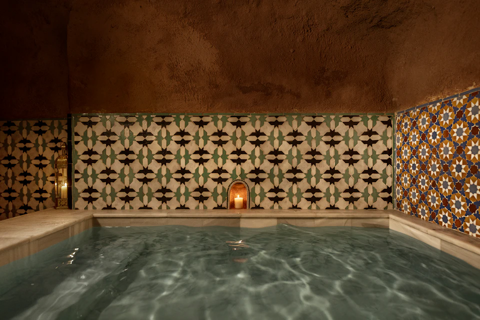 A tiled indoor pool with patterned walls, a small niche holding a lit candle, and a water reflection.
