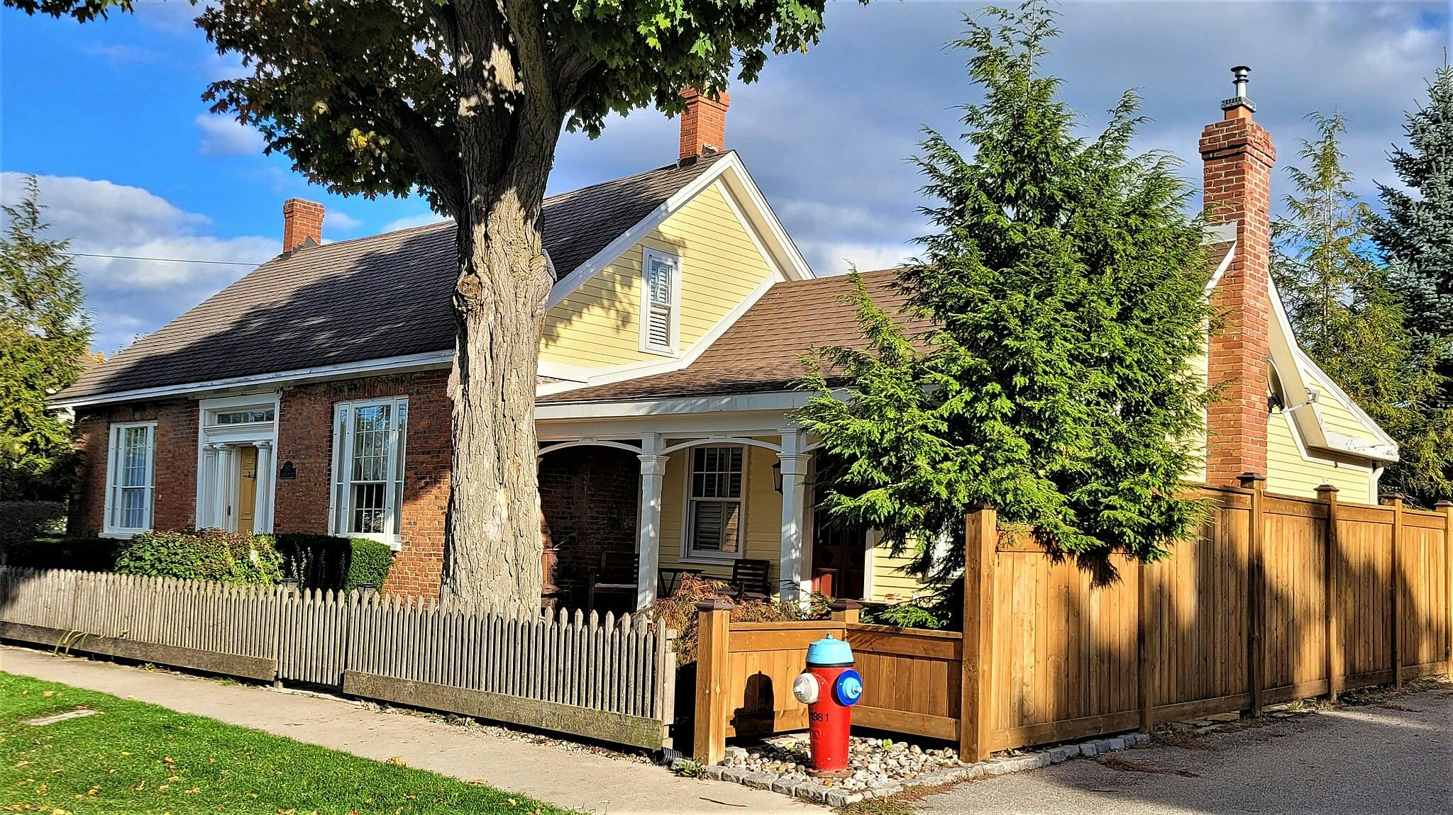 벽돌과 노란색 외관, 흰색 테두리, 피켓 울타리, 큰 나무, 빨간색 소화전을 갖춘 2층 주택입니다.