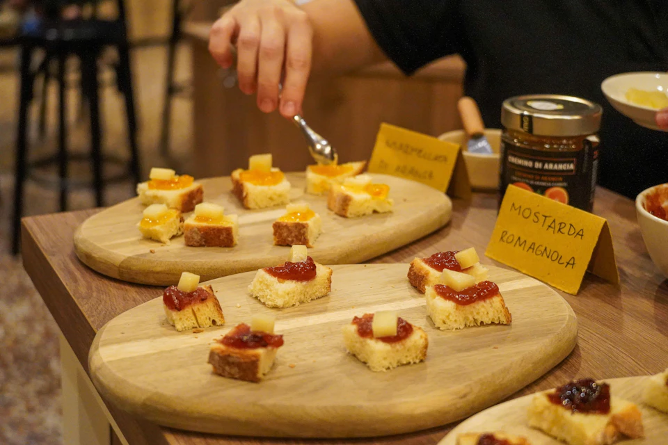 Hand reaching for bite-sized bread pieces topped with various jams and cheese cubes on wooden boards, with yellow labels in the background.