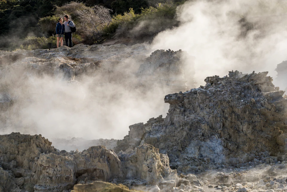 Two people stand near steaming geothermal rock formations, surrounded by vegetation.
