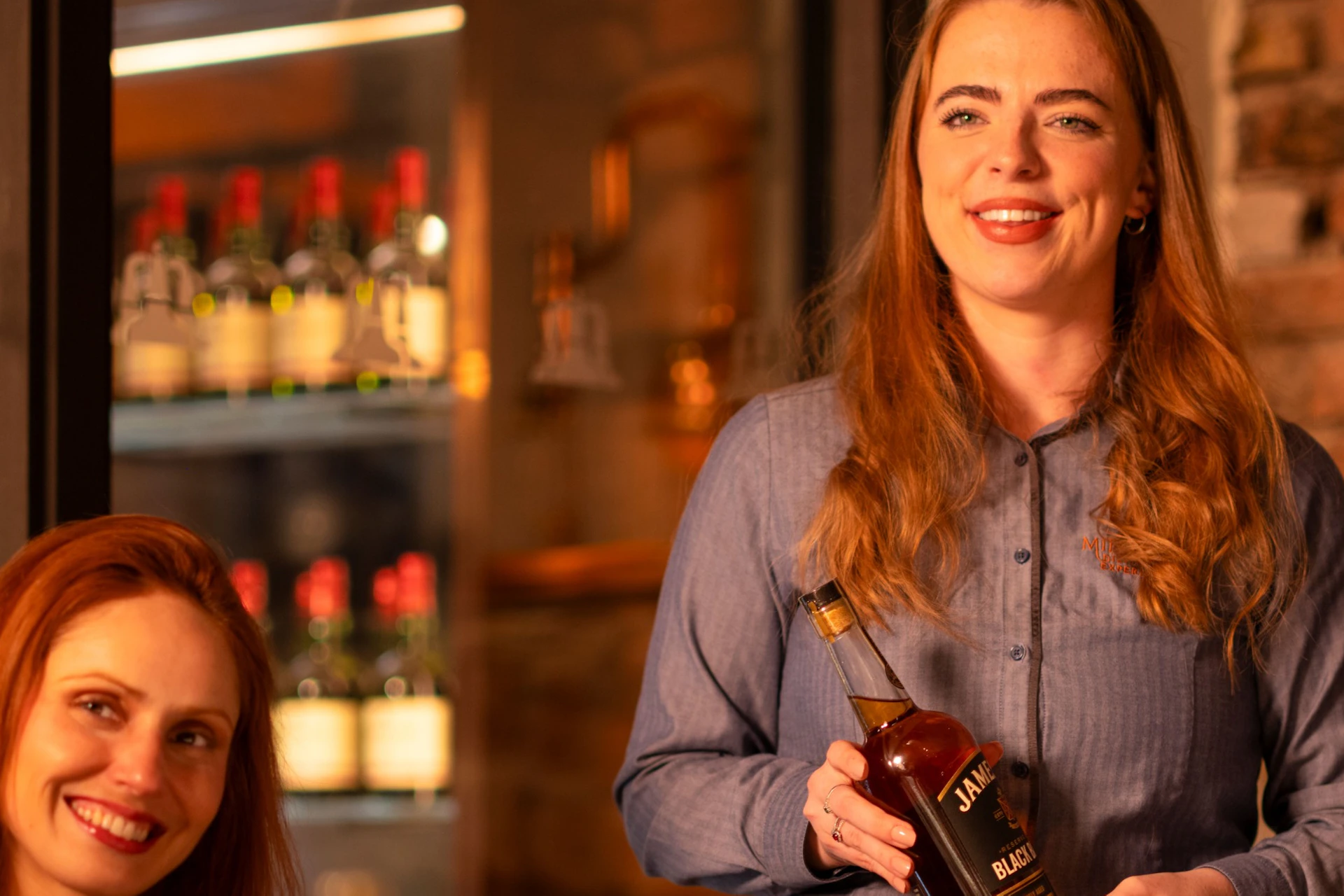 A woman presenting a bottle at a table with whiskey glasses, with another woman smiling next to her in a cozy bar setting.