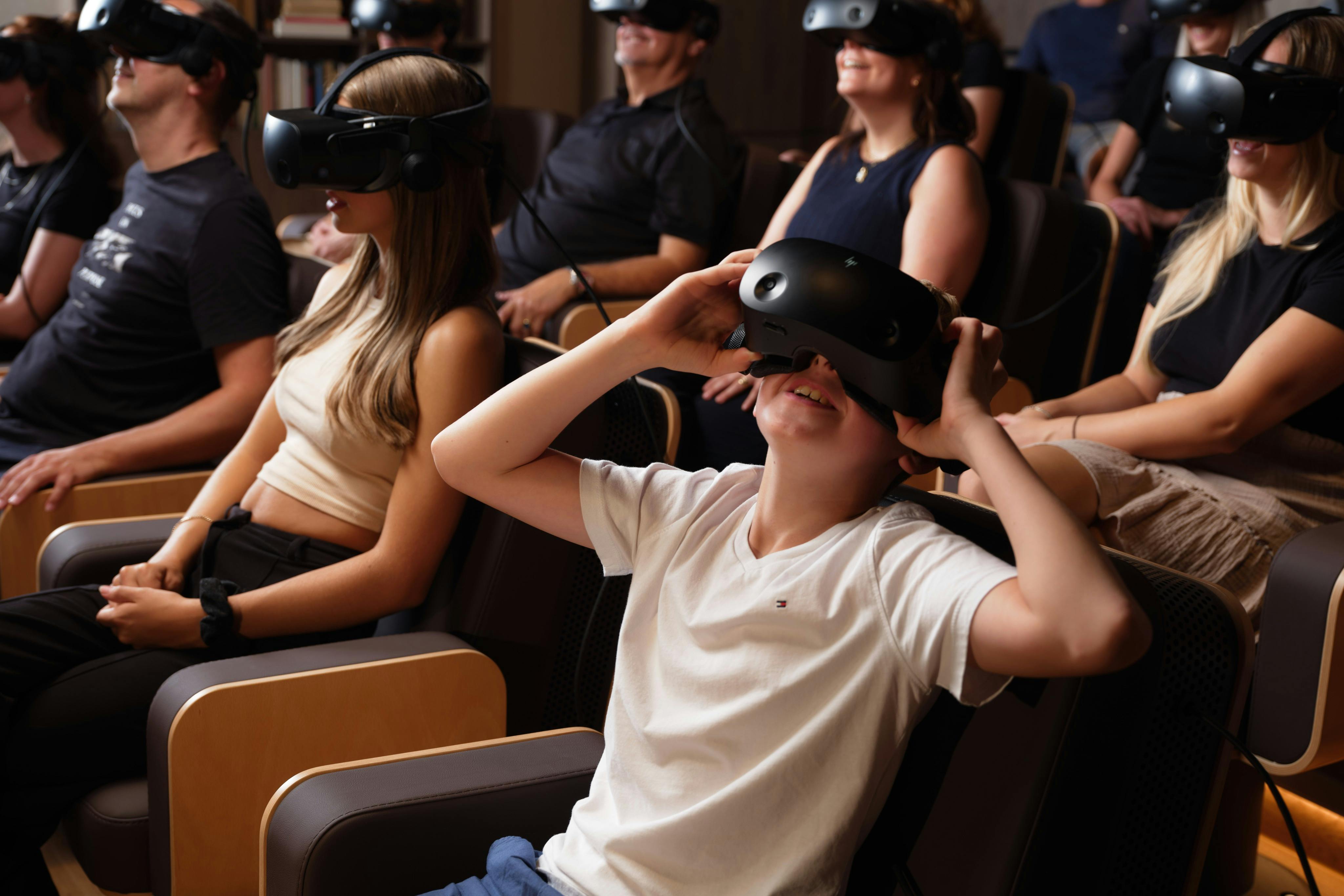 En grupp människor som sitter i ett rum, klädda i virtual reality-headset, med ett barn i förgrunden som håller i headsetet.