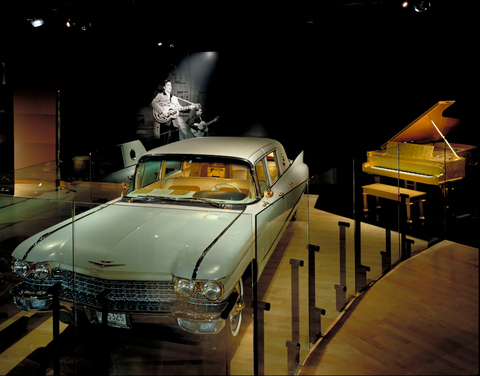 Exhibit featuring a vintage car, a grand piano to the right, and a photo of a musician with a guitar in the background.