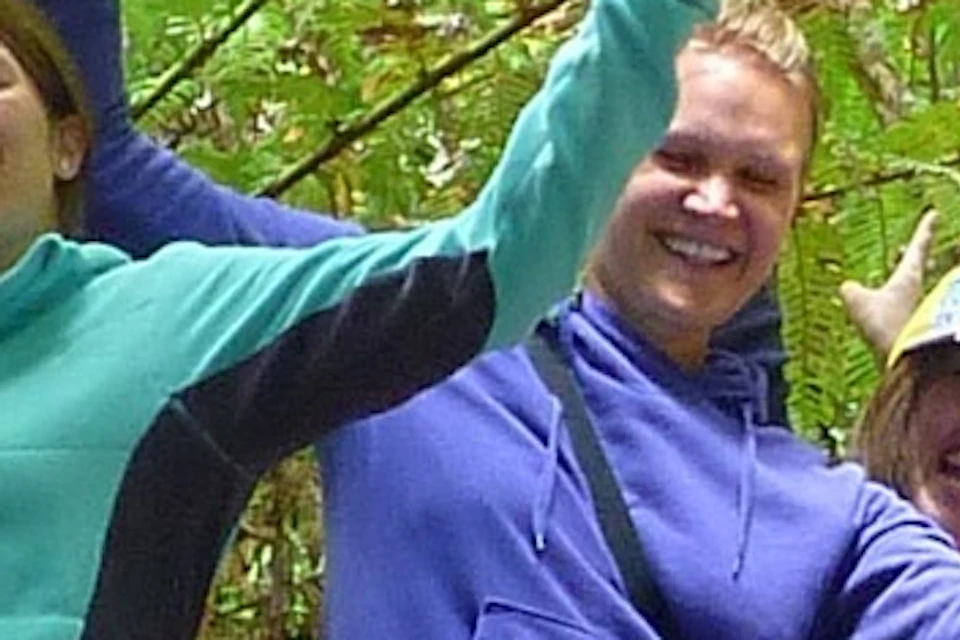 A group of seven people stand on a large fallen tree trunk in a lush forest, smiling and raising their arms under tall green ferns.