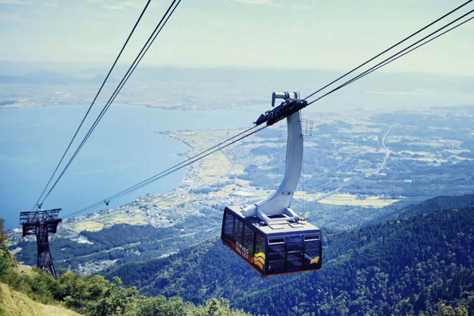 A cable car suspended over a mountainous landscape with a lake and a town visible in the background.