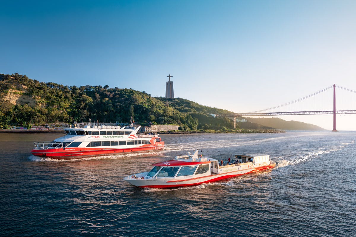 붉은 보트와 흰 보트 두 척이 강을 따라 항해하고 있습니다. 오른쪽에 다리가 있는 언덕을 배경으로 높은 동상이 서 있습니다.