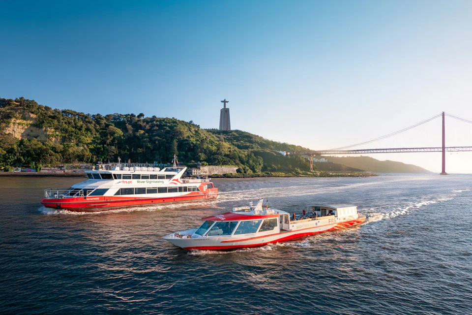 Two red and white boats are cruising on a river. A tall statue stands on a hill in the background with a bridge on the right.