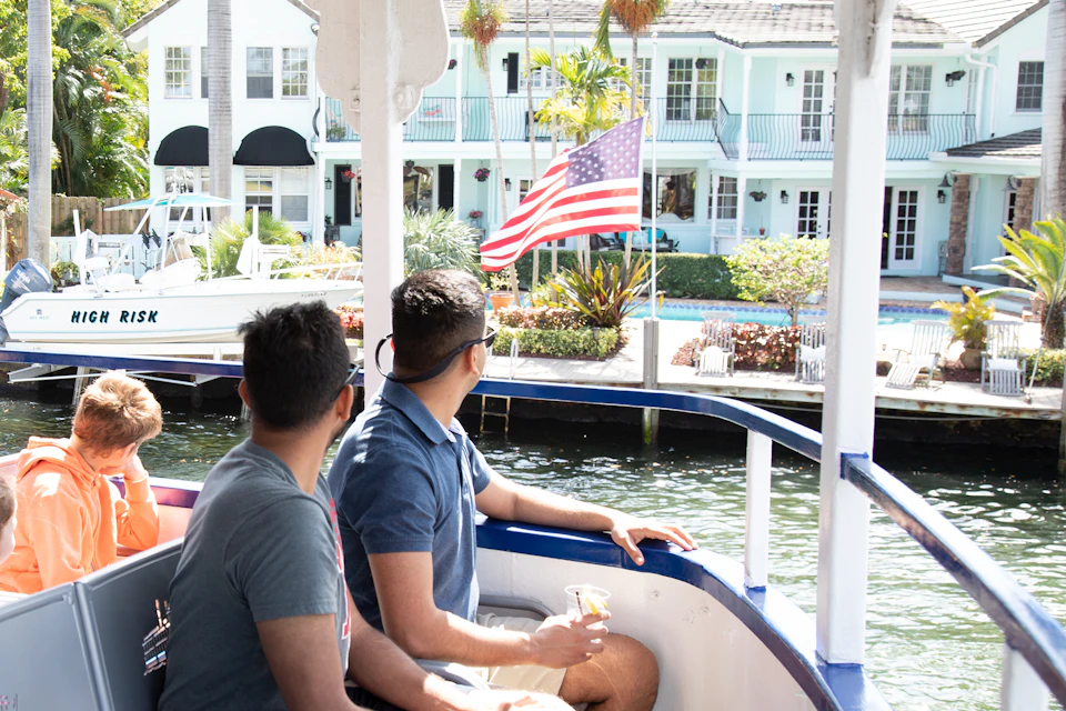 Two men on a boat look towards an American flag and pastel-colored waterfront houses with balconies and lush greenery.