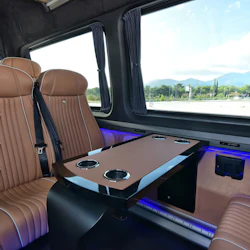 Lyxig fordonsinteriör med solbrända lädersäten, ett bord med mugghållare och ett stort fönster som visar en vacker utsikt utomhus.