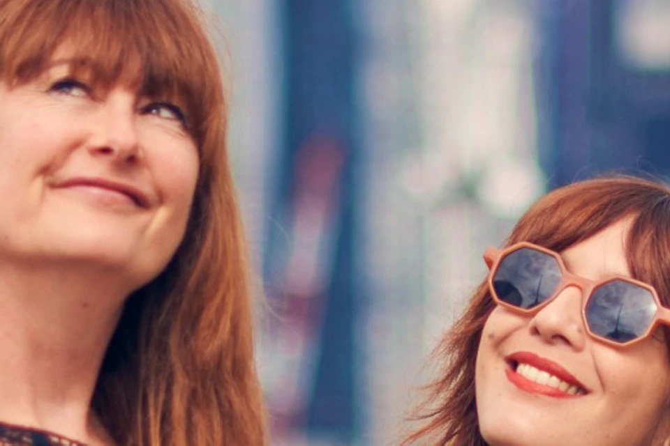 Two women taking a selfie with a city skyline in the background on a sunny day. One woman is holding up a phone.
