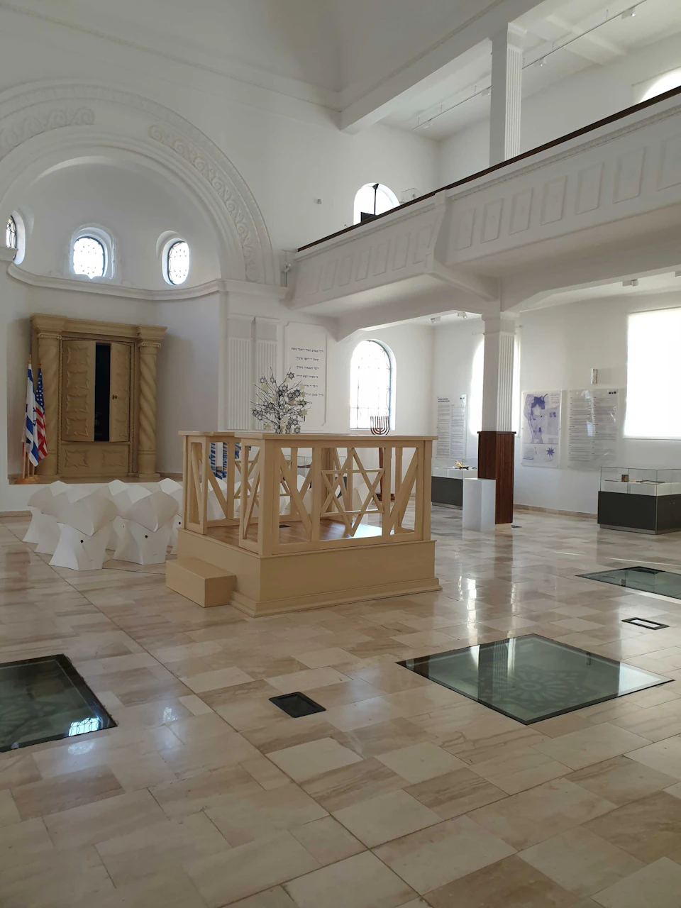 A bright, spacious room with marble floor, a central wooden railing structure, white display panels, and arched windows.