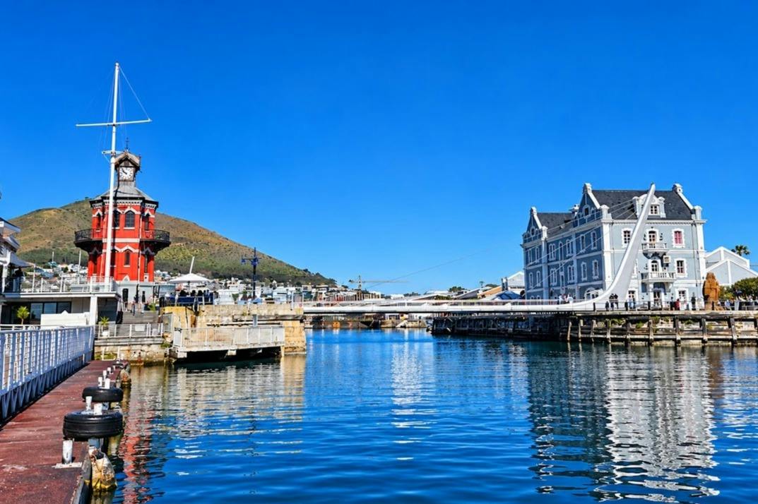 A calm waterfront scene with red and white buildings, a bridge, and a mountain in the background under a clear blue sky.