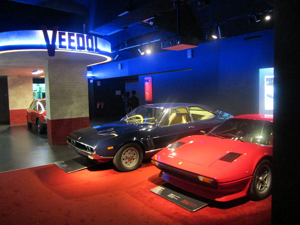 Two vintage sports cars, one blue and one red, displayed in a dimly lit showroom with a "VEEDOL" sign above.