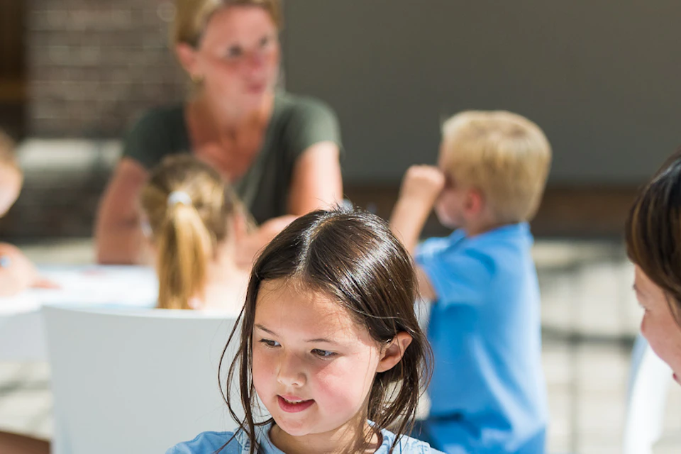 Children and adults seated at white tables, engaging in drawing activities with colored pencils, in a sunlit indoor setting.