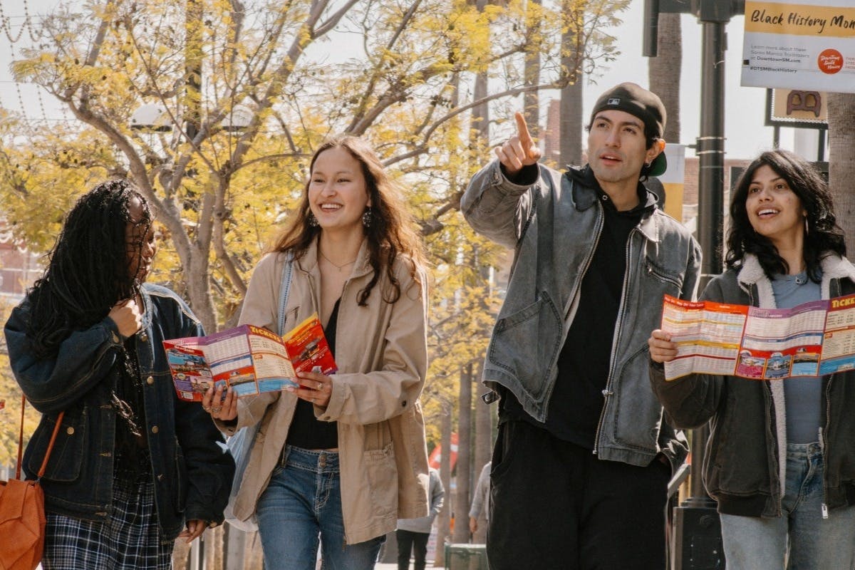 Three friends walking outdoors on a sunny day, holding a map and conversing, with trees and a bright background visible.