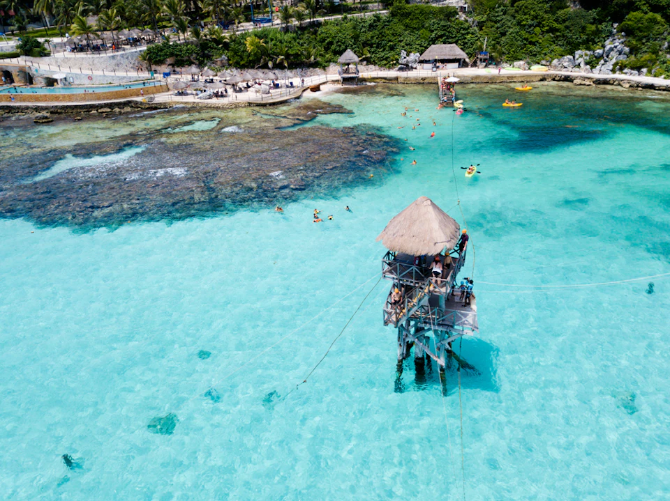 Aerial view of a tropical shoreline with clear turquoise water, a thatched-roof lookout tower, and scattered swimmers and kayakers.