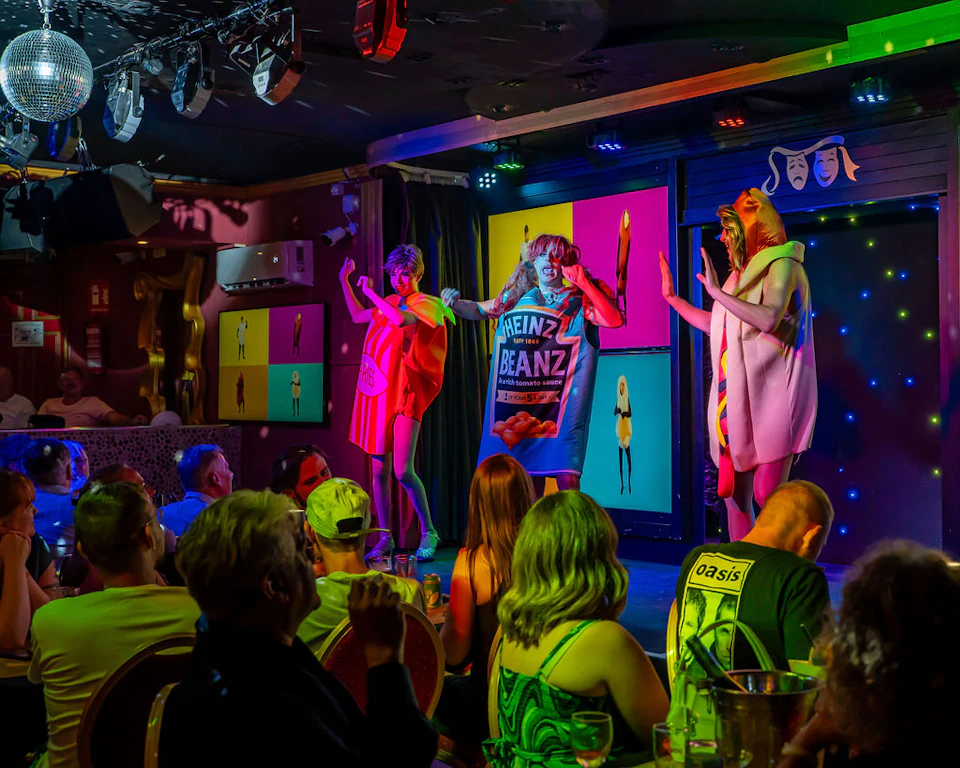 Three performers in colorful costumes, including food-themed outfits, entertain an audience in a vibrant stage setting.