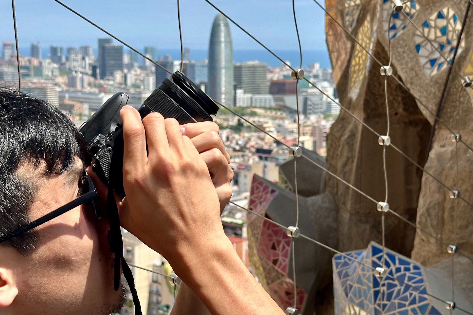 Person holding a camera, photographing a city skyline with tall buildings and a unique, cylindrical skyscraper in the background.