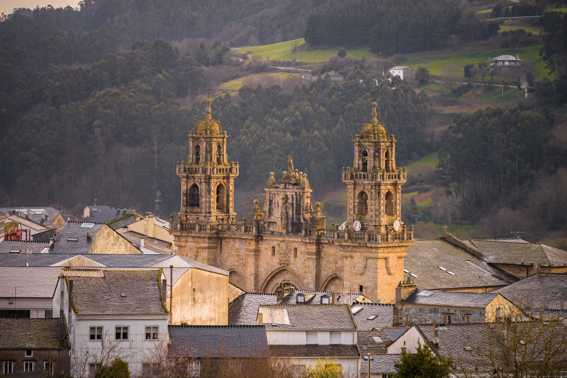 A historic stone cathedral with two bell towers, situated in a town surrounded by lush green hills and forests.