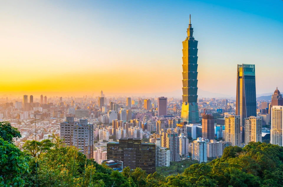 Cityscape at sunset with a tall, uniquely designed skyscraper surrounded by numerous buildings and greenery in the foreground.