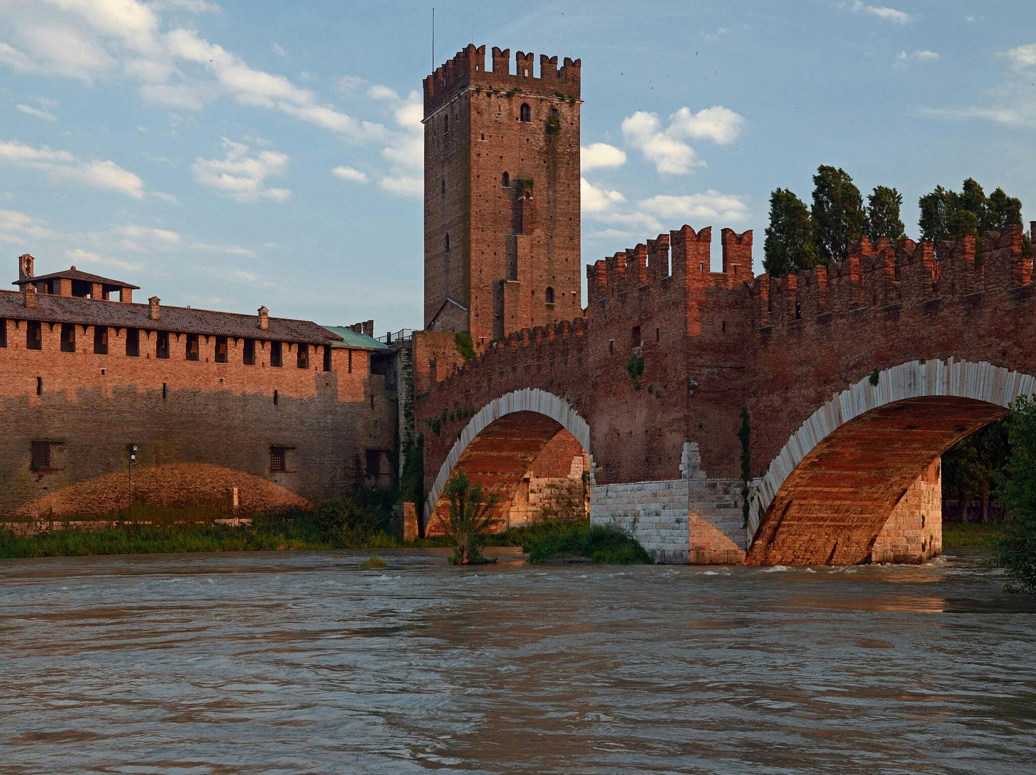 Old brick bridge with arches spans a wide river; connecting to a walled fortress with a tall tower at sunset.