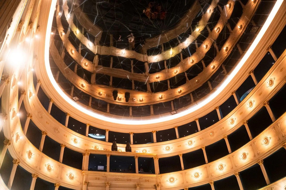 Interior view of a theater with multiple balconies and warm lighting, reflected in a mirrored ceiling.