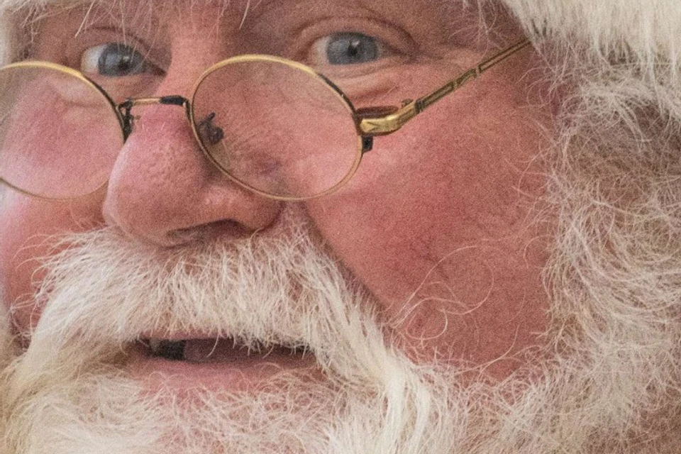 An elderly man dressed in a Santa outfit with a white beard, wearing glasses, sits by a window holding a coffee cup on a boat.