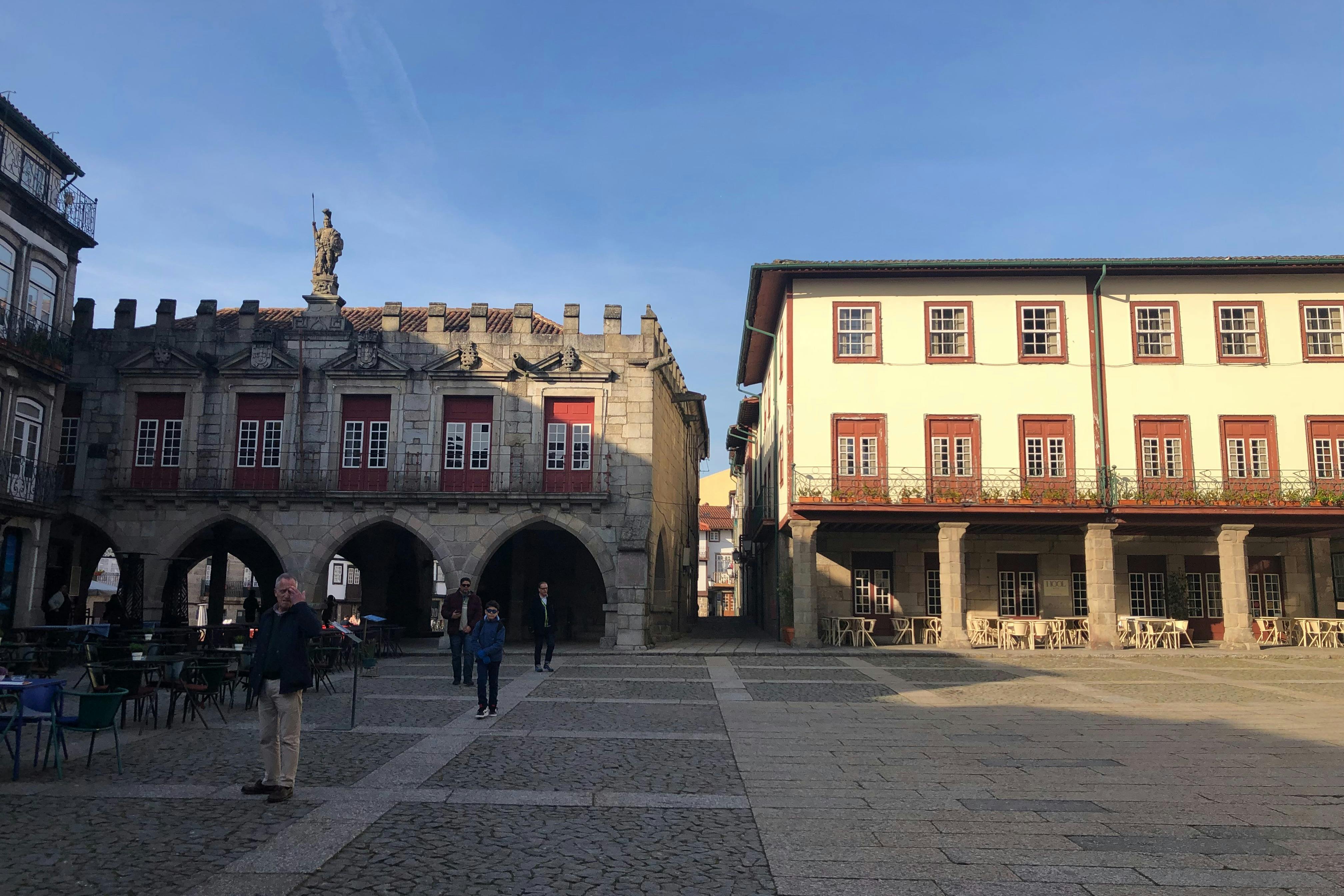 A town square with a historic building featuring arches on the left and a two-story building with red window frames on the right. Few people present.