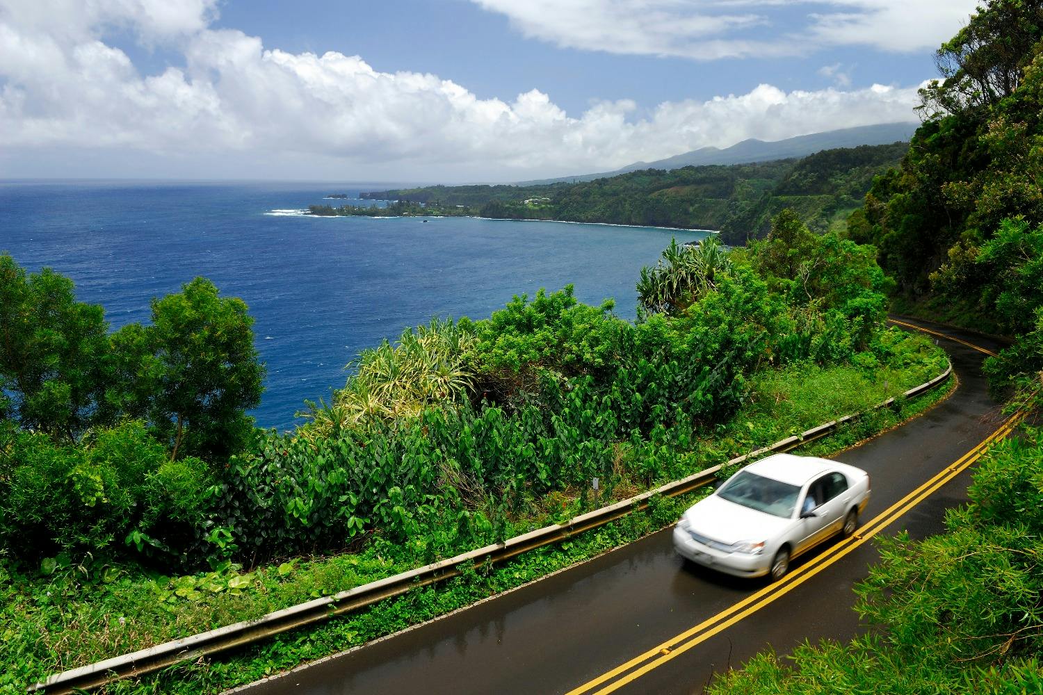 A white car drives on a coastal road surrounded by lush green vegetation, overlooking a blue ocean under a partly cloudy sky.