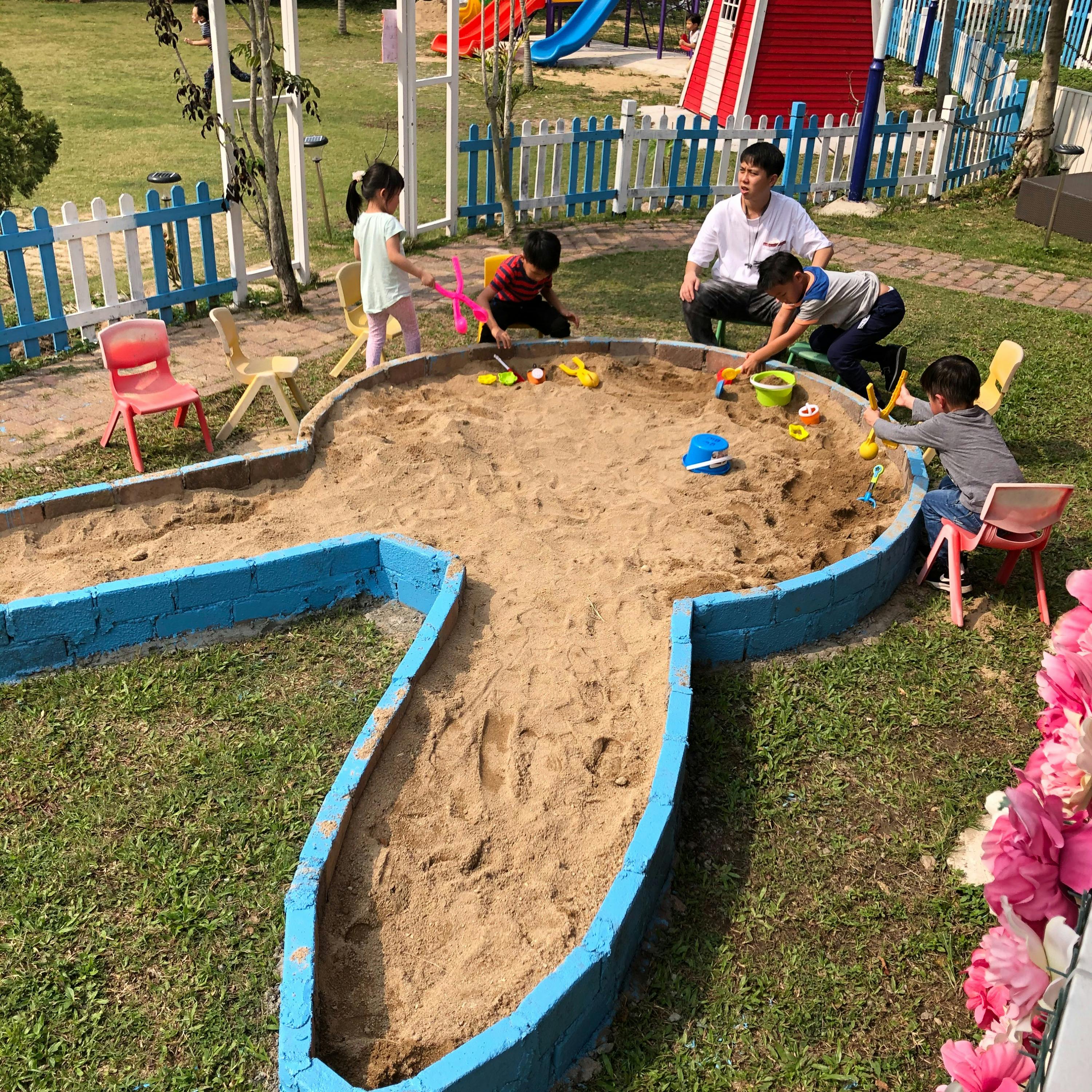 Children and an adult playing in a sandpit shaped like a rabbit, surrounded by grass, colorful chairs, and a playground.