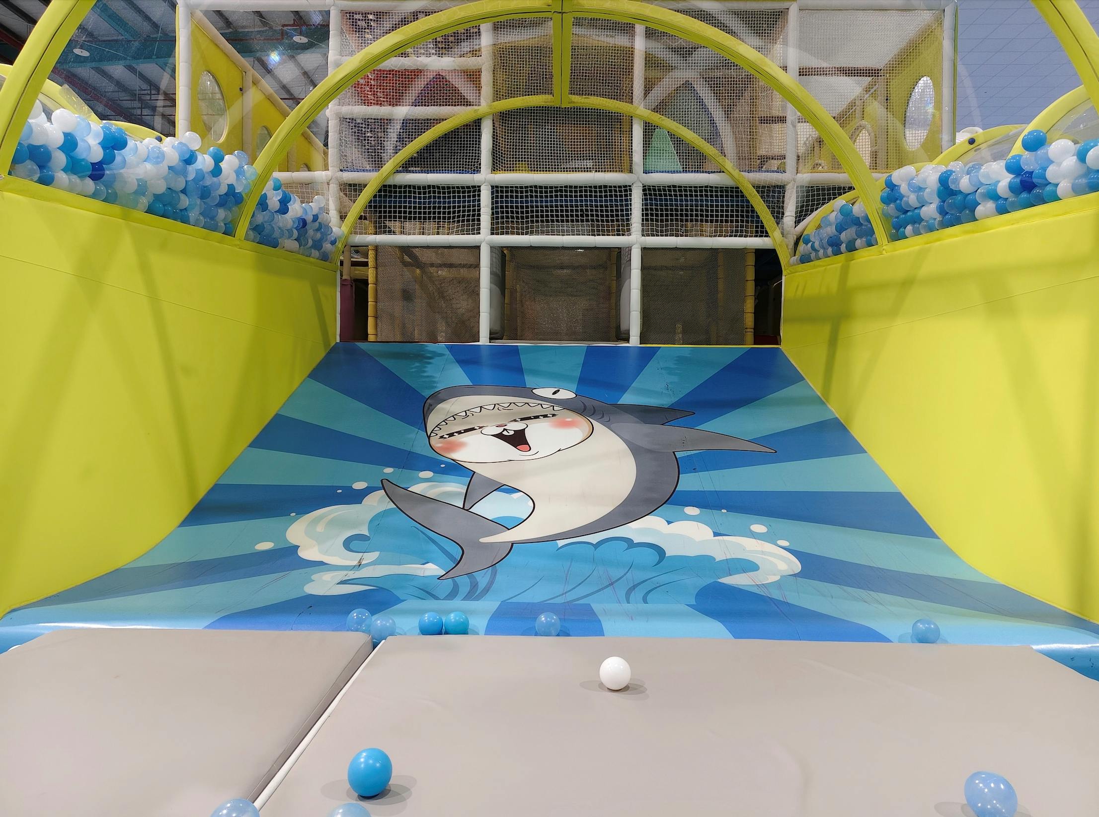 Indoor play area with a shark illustration on a slide featuring blue and white balls and yellow barriers.