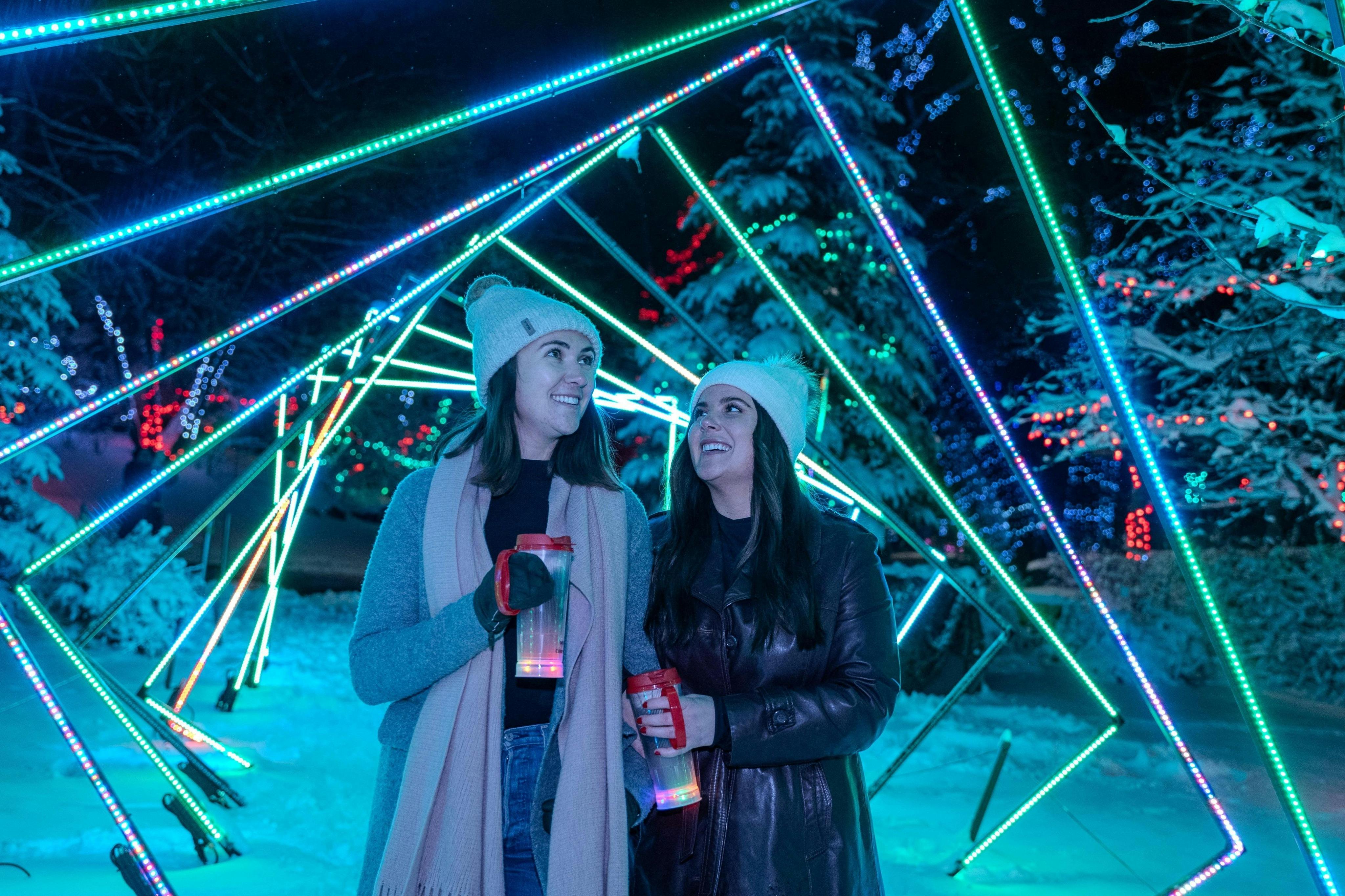 겨울 옷을 입은 두 명의 여성이 다채롭고 기하학적인 조명 조각이 있는 눈 덮인 지역에 서서 미소를 짓고 조명 컵을 들고 있습니다.