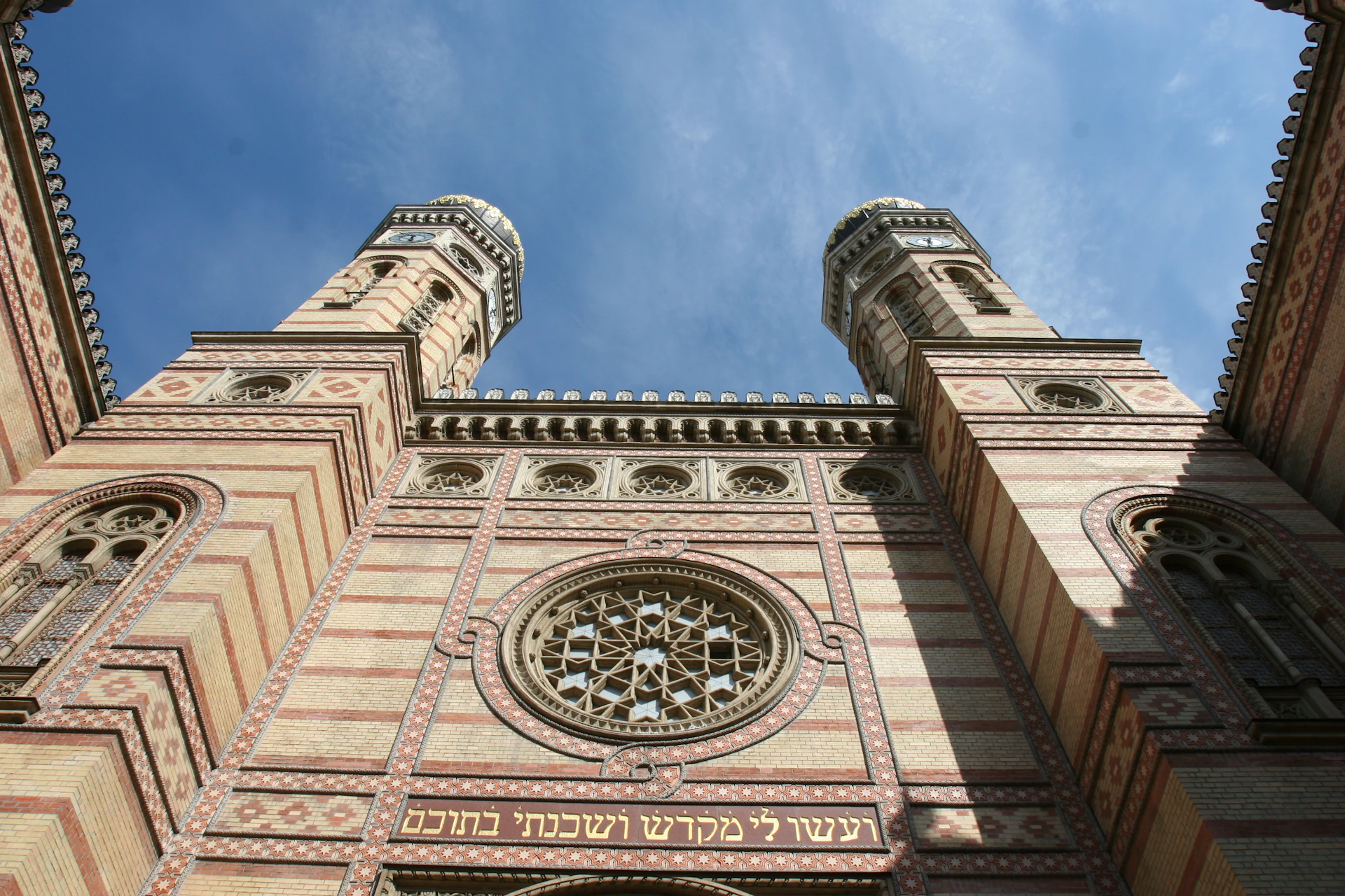 Ornate building facade with two tall towers, a circular stained glass window, and decorative Hebrew inscriptions, under a blue sky.