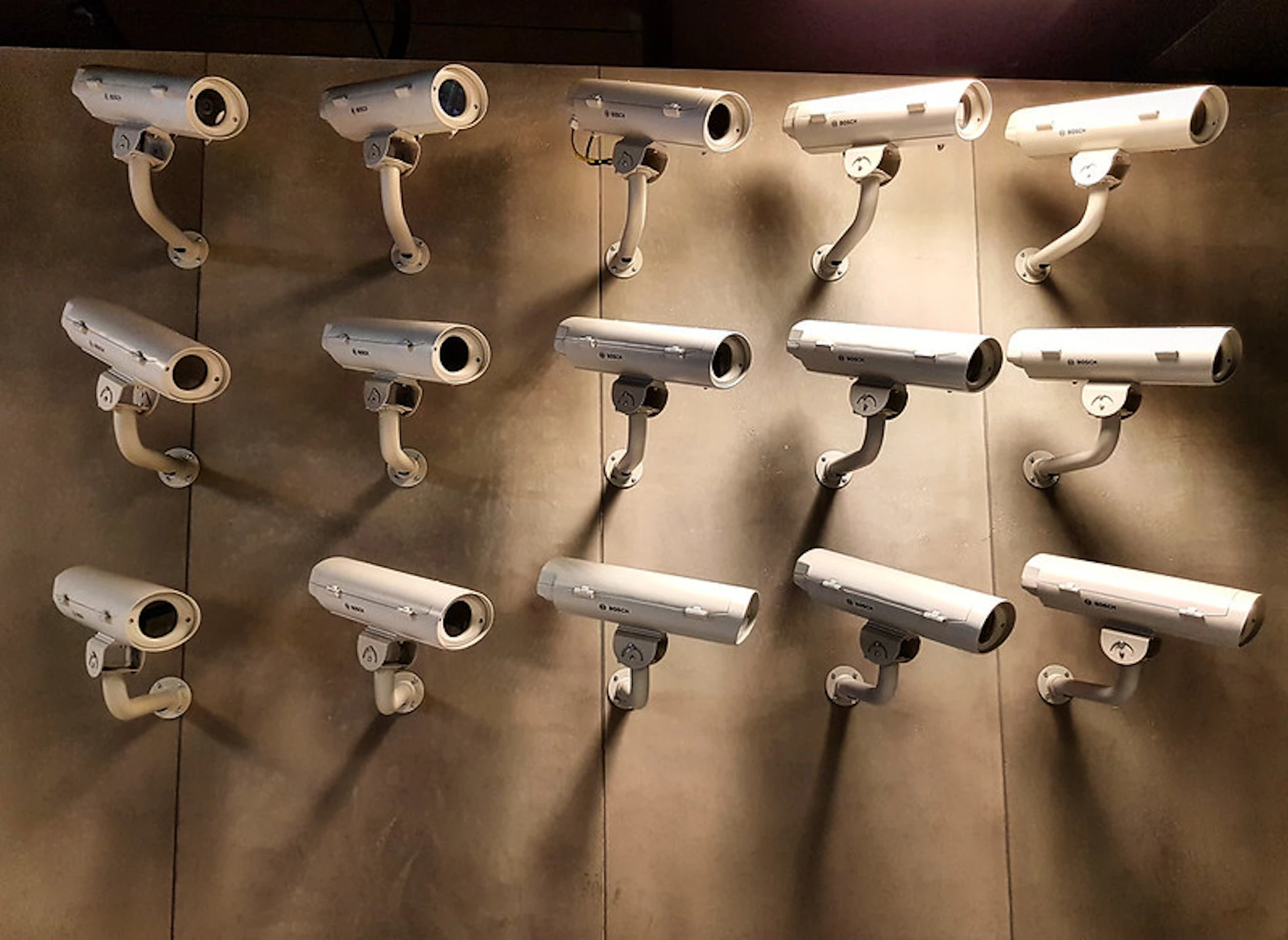 Multiple white security cameras are mounted on a beige wall in a grid pattern, all pointing in various directions.