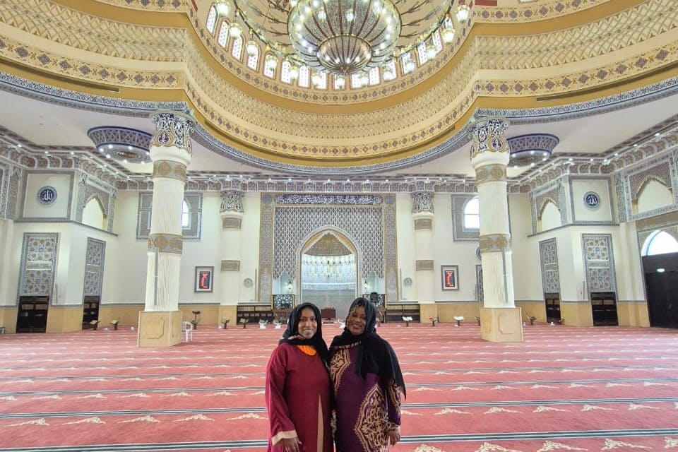 Deux femmes en tenue traditionnelle se tiennent à l'intérieur d'une grande mosquée aux motifs complexes, aux grandes colonnes et au plafond en forme de dôme.
