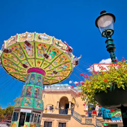 En färgglad svingtur i en nöjespark, med dekorativa paneler, en gatlykta och en blommig planter under en blå himmel.
