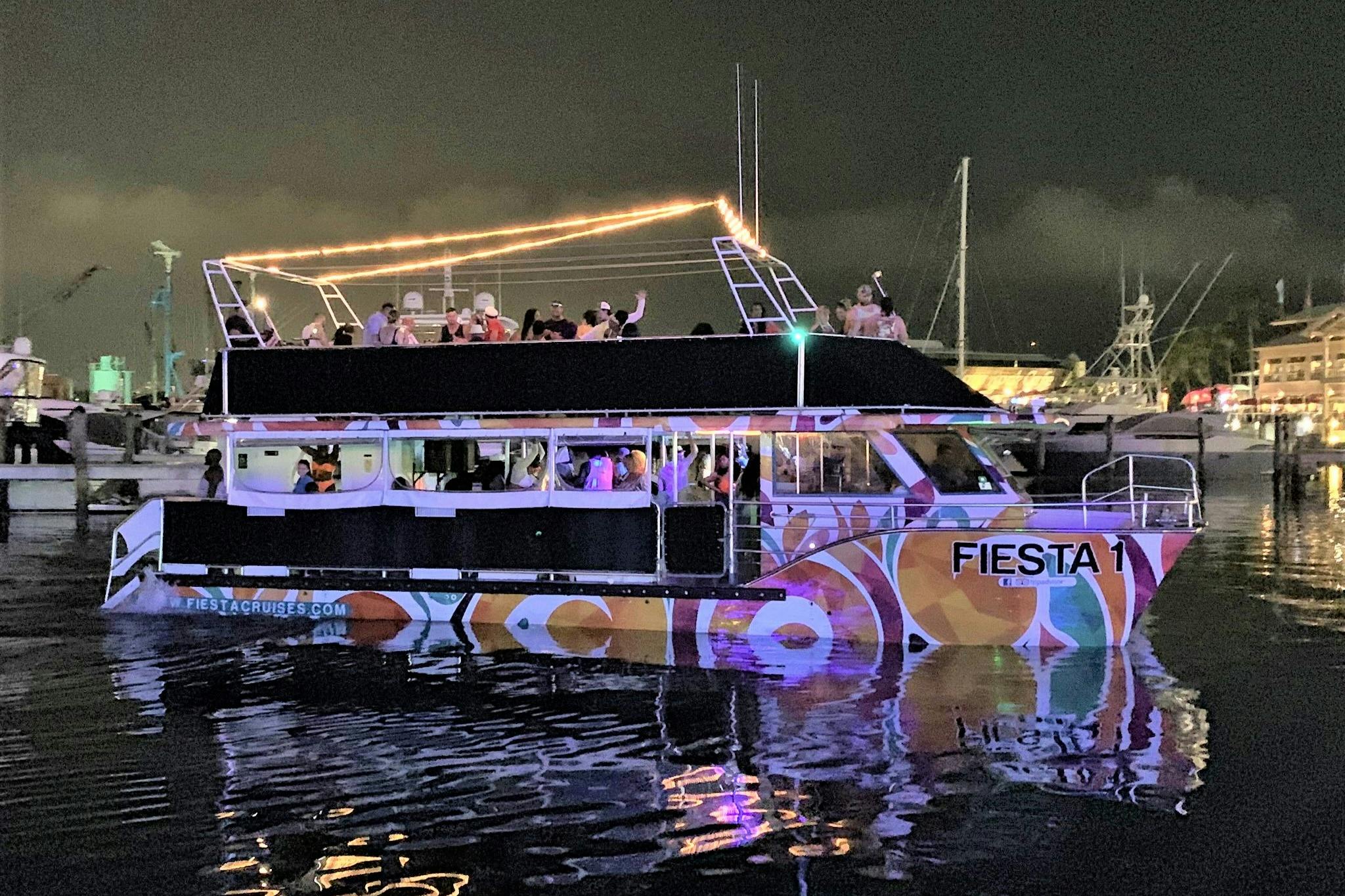 A party boat with colorful patterns and people on the upper deck at night, illuminated by lights, floating on the water.