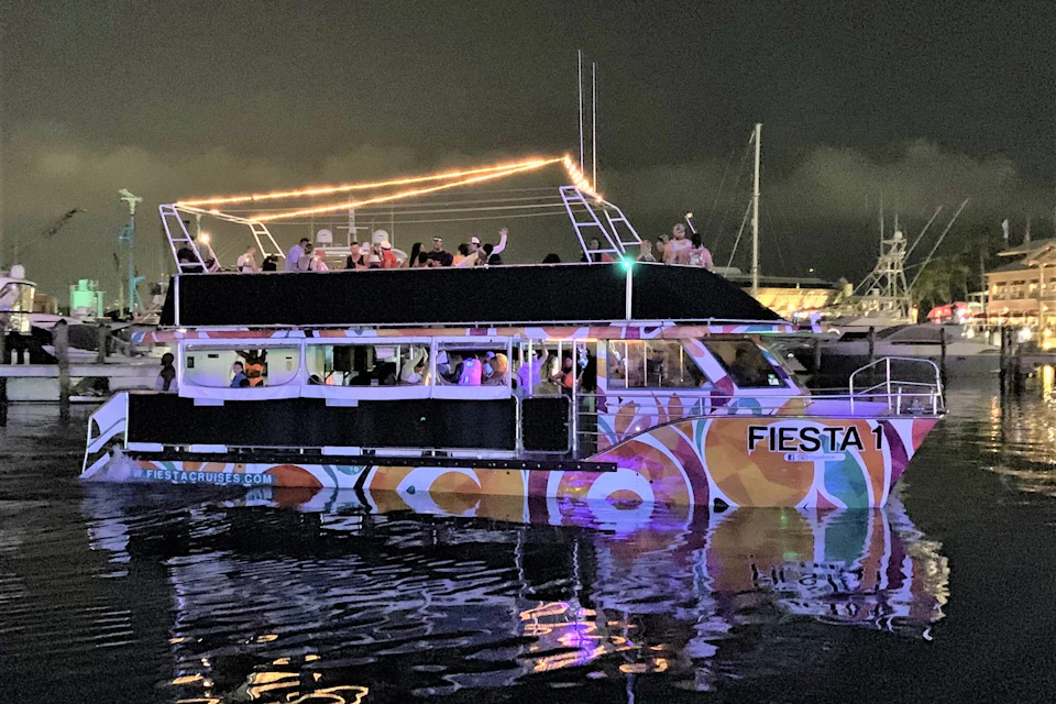 A party boat with colorful patterns and people on the upper deck at night, illuminated by lights, floating on the water.