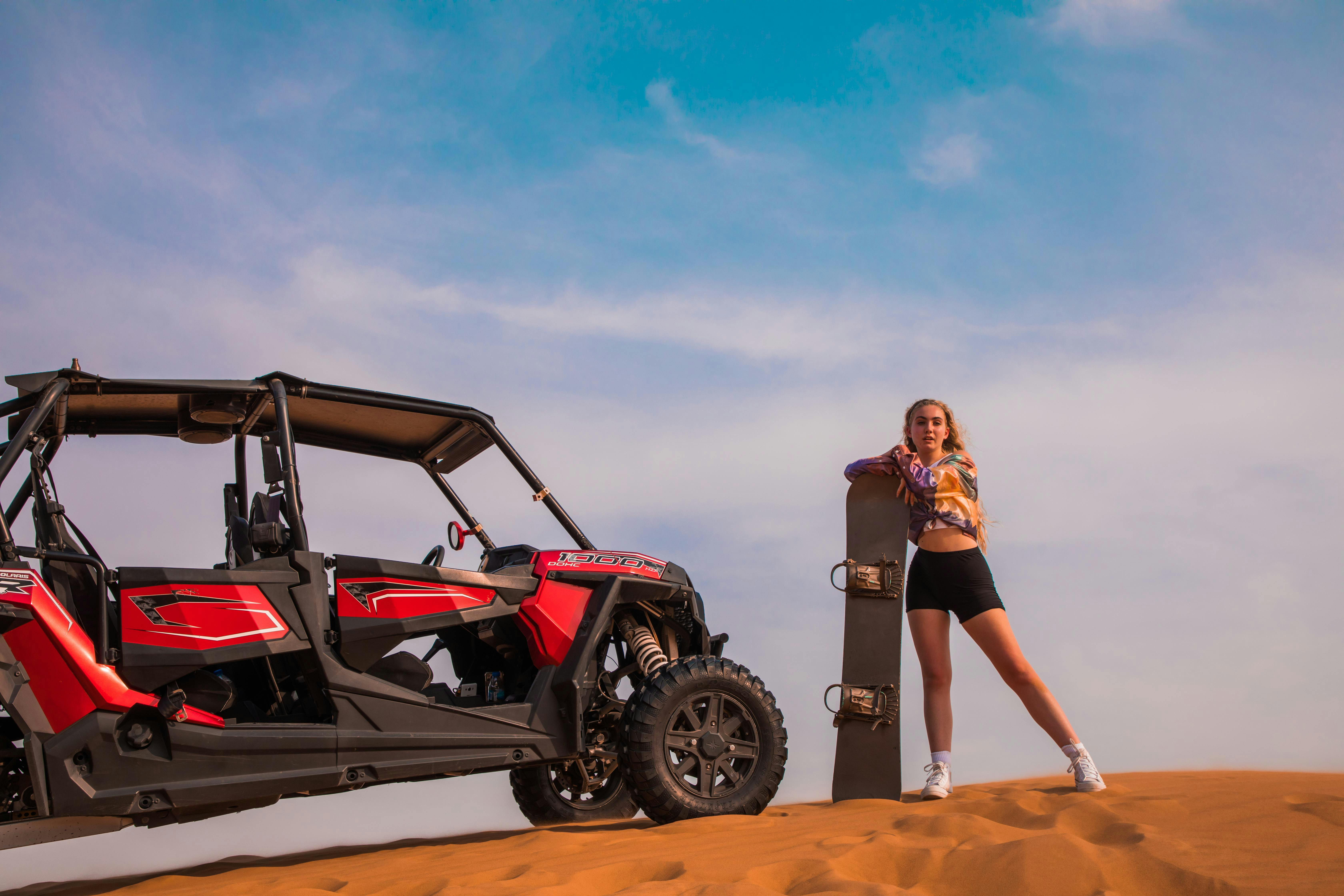 Una donna si trova nel deserto accanto a un dune buggy rosso, con in mano una tavola da sabbia, sotto un cielo blu con nuvole.