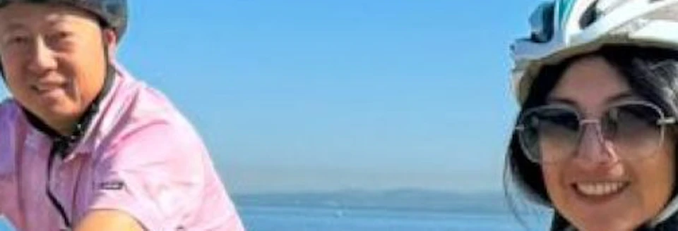 Two people riding bicycles on a sandy beach under a clear blue sky, wearing helmets and smiling.