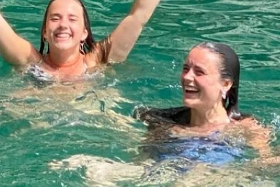 Three people swimming in clear green water, smiling and raising their arms in a gesture of joy.