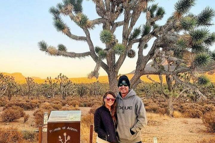 Two people stand smiling in front of a Joshua tree in a desert landscape during daylight.
