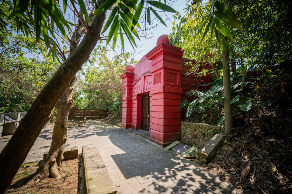 A red building with an arched doorway surrounded by trees and greenery on a sunny day.
