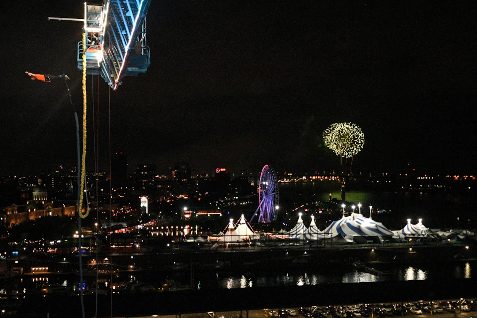 A nighttime cityscape showing a lit Ferris wheel, circus tents, fireworks, and a crane arm overhead.