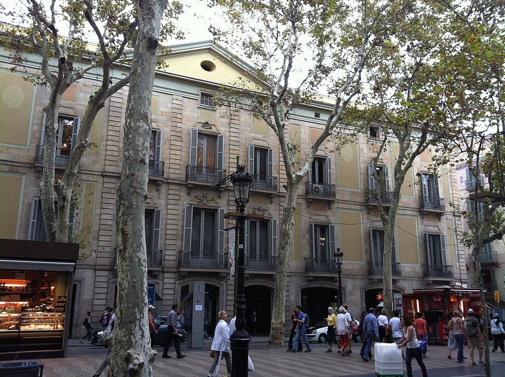 Lidé procházejí kolem staré vícepodlažní budovy s velkými stromy a pouliční lampou před sebou. Vlevo je malý stánek s občerstvením.
