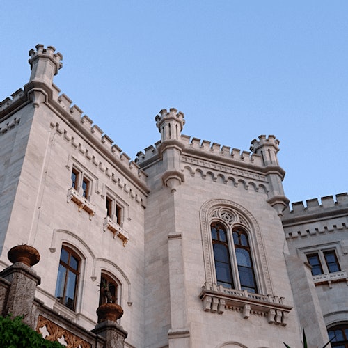 Stone castle facade with arched windows and decorative towers against a clear blue sky.